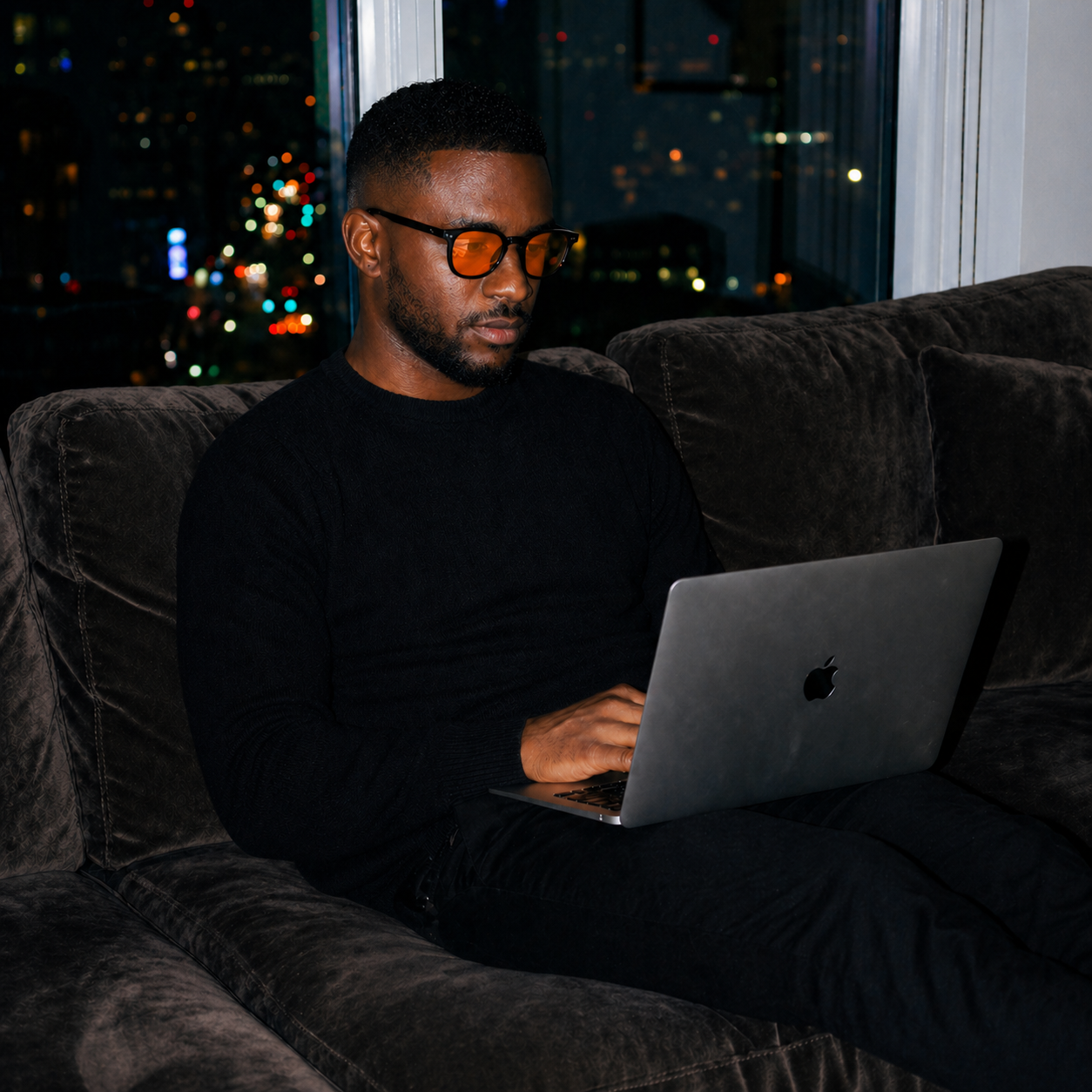 Man wearing NEURIX™ on couch with laptop at night, city lights behind.